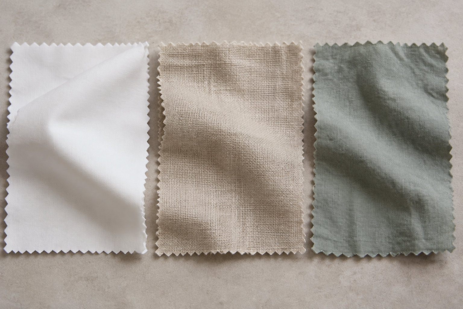 Trois échantillons de tissu blanc, beige et vert grisé sont posés côte à côte sur une surface beige.